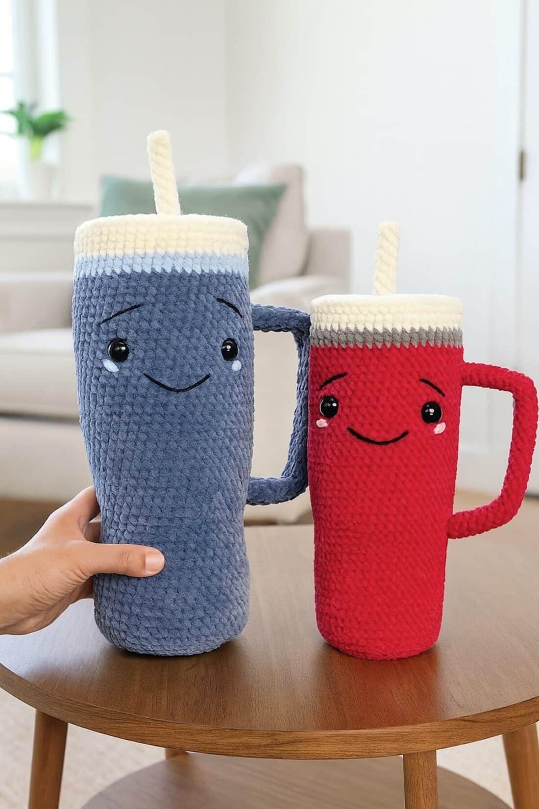 Two Amigurumi tumblers, one dark gray and one dark pink, sitting on a wooden coffee table in a cozy living room.