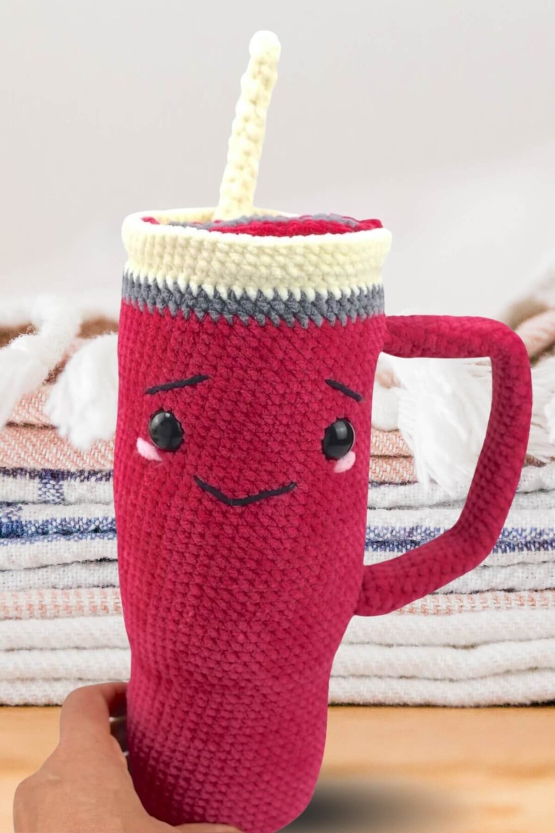 A hand holding a pink Amigurumi tumbler, sitting on a white surface with flowers in the background.