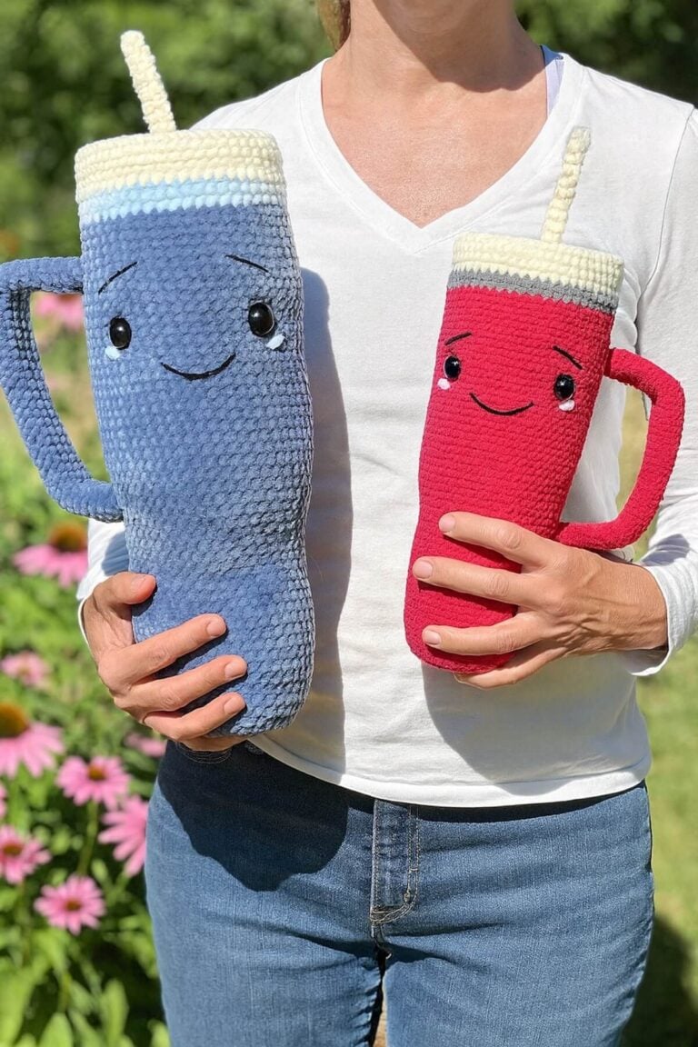 A person holding two crochet Amigurumi tumblers, one jumbo gray and one hot pink, both with smiling faces.