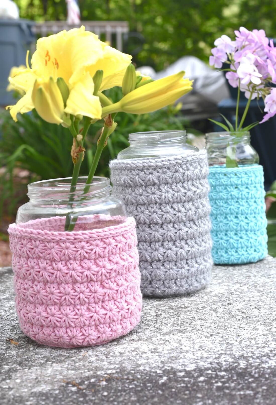 Three glass jars on pink, blue, and gray crochet sleeves displayed outdoors with flowers in them.