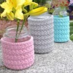 Three mason jars on pink, blue, and gray crochet covers displayed outdoors with flowers in them.
