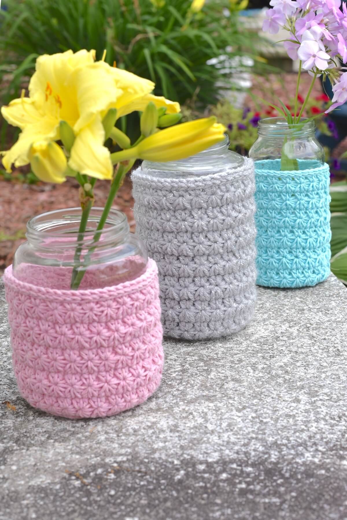 Three mason jars on pink, blue, and gray crochet covers displayed outdoors with flowers in them.