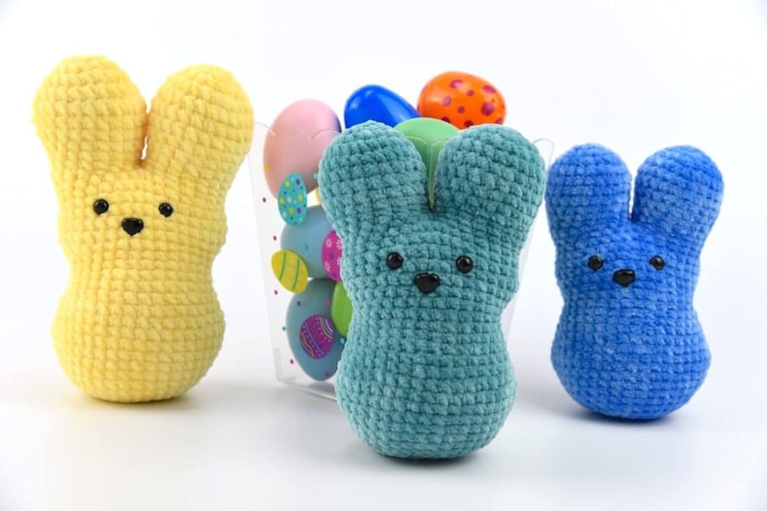 Three crochet peep bunny plushies in yellow, teal, and blue yarn, made with velvet yarn and shaped like marshmallow Easter treats, displayed in front of a white background.