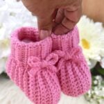 A hand holding a pair of pink crochet baby booties with a white fur fabric in the background.