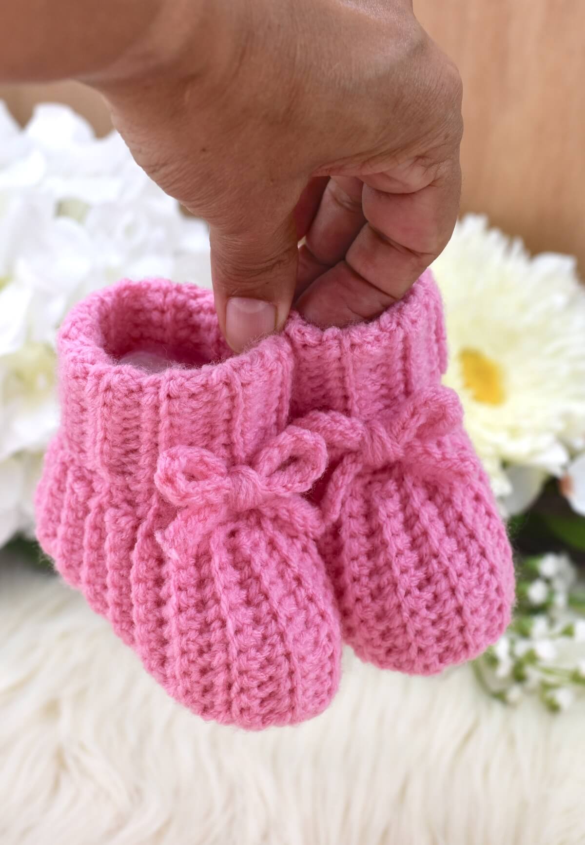 A hand holding a pair of pink crochet baby booties with a white fur fabric in the background.