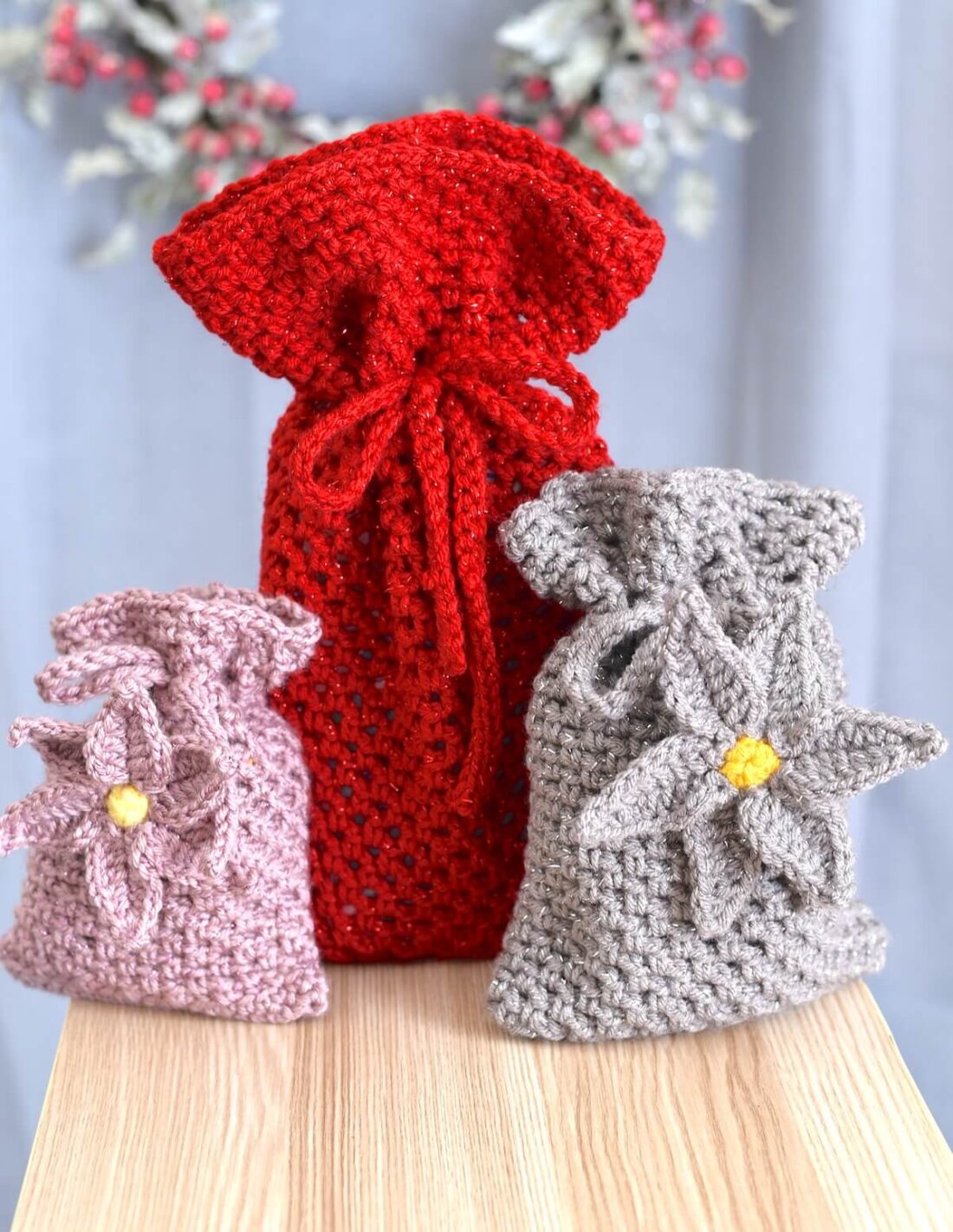 Set of three crochet gift bags in red, pink, and gray displayed on a wooden surface.