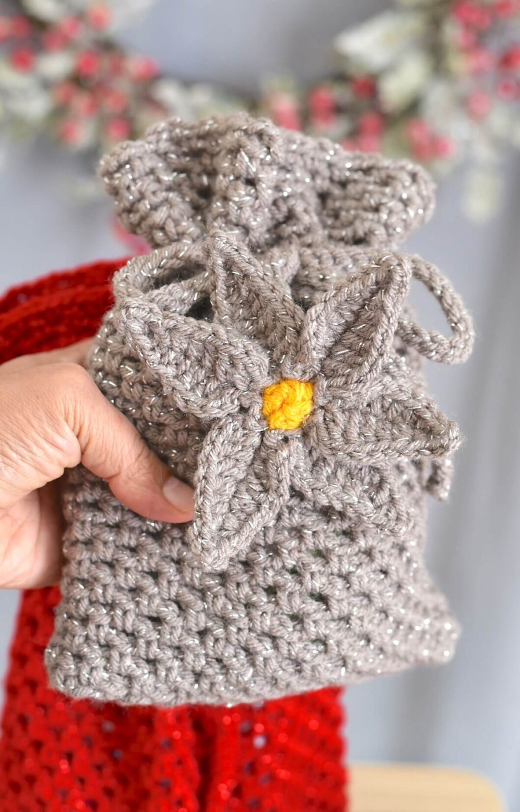 A hand holding a medium size, gray crochet gift bag decorated with a crochet flower.