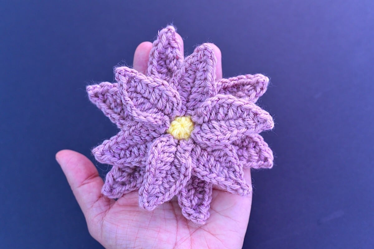 A crochet poinsettia flower with layered pink petals and a yellow center layered flat on a palm of a hand.