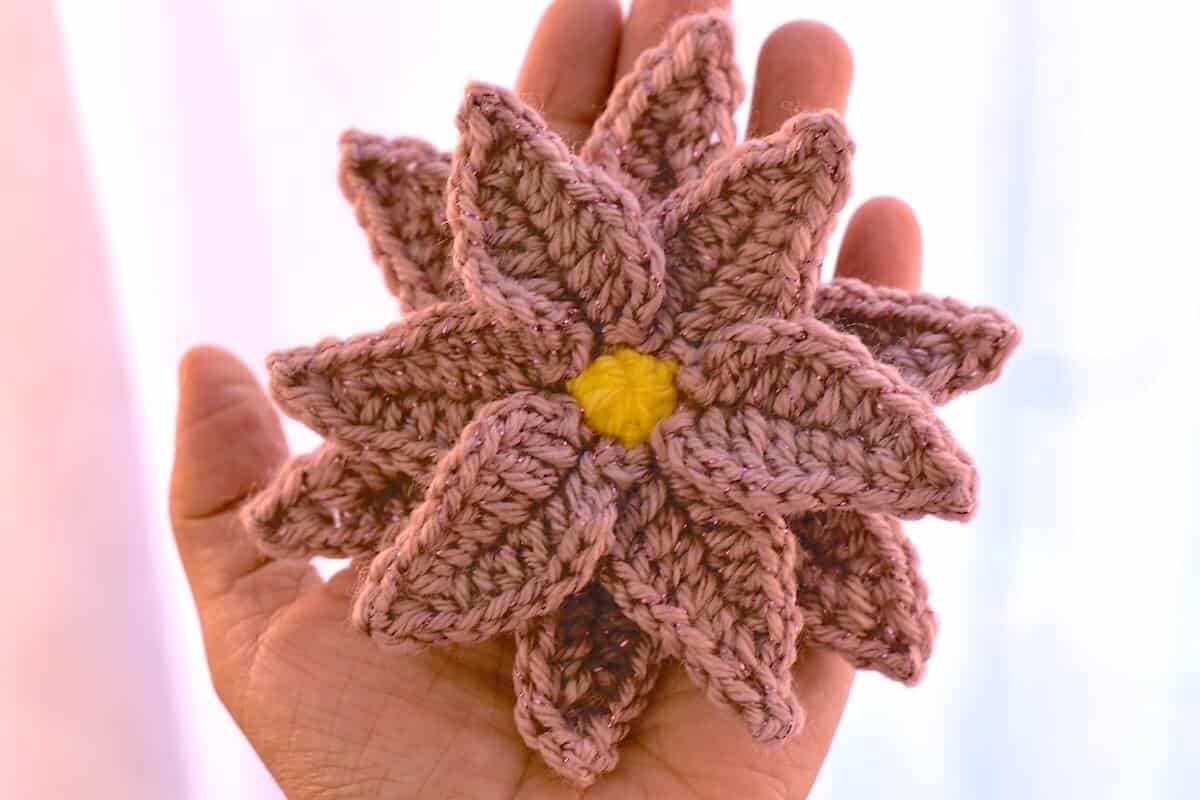 A crochet poinsettia flower with layered pink petals and a yellow center displayed on a palm of a hand.