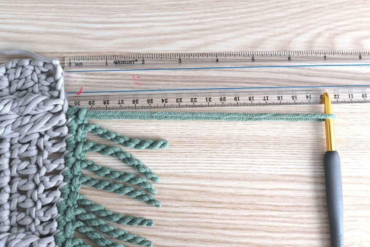 Close-up of an unfinished croche swatch, a half-finished finge with green yarn, and a hook, showing how to make a twisted fringe.