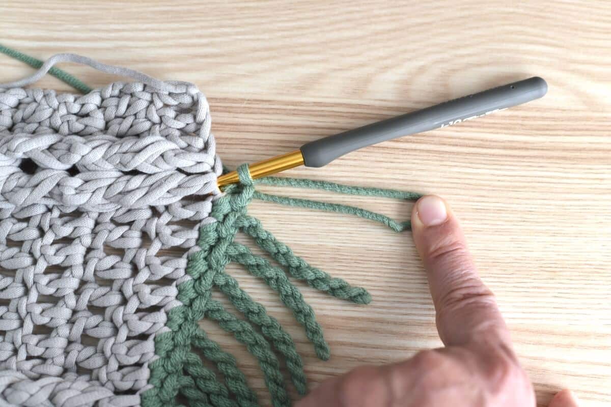 A close-up of a hand and a brown crochet swatch, showing a twisted fringe made with green yarn for crochet blankets.