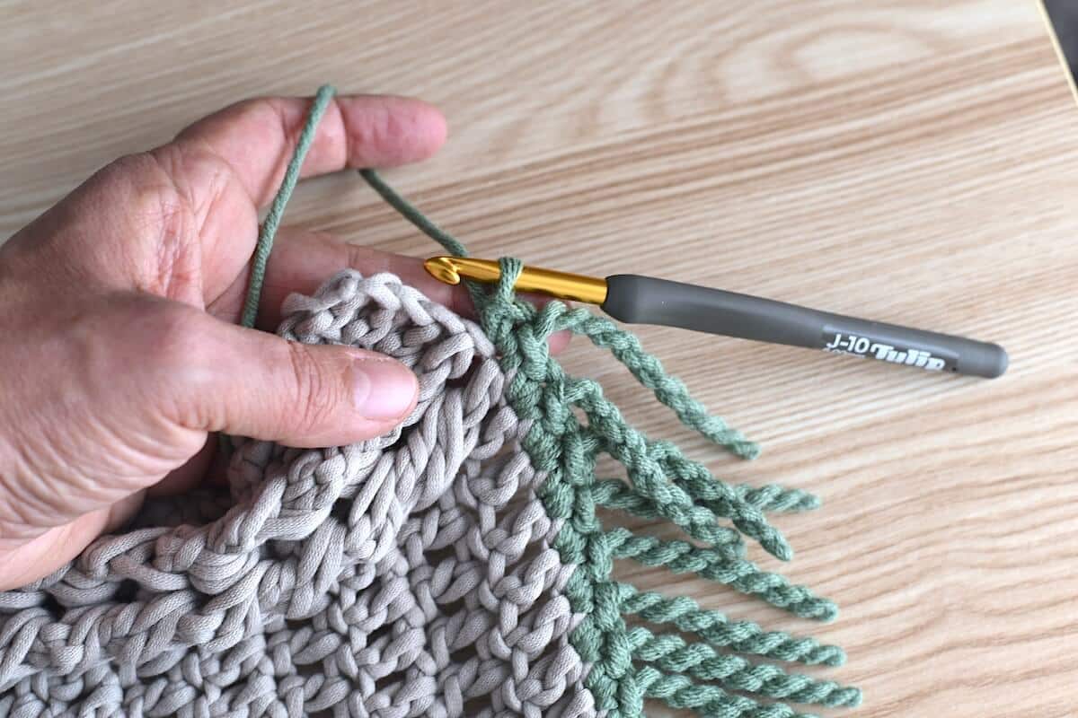 A close-up of a hand and a brown crochet swatch, showing a twisted fringe made with green yarn for crochet blankets.