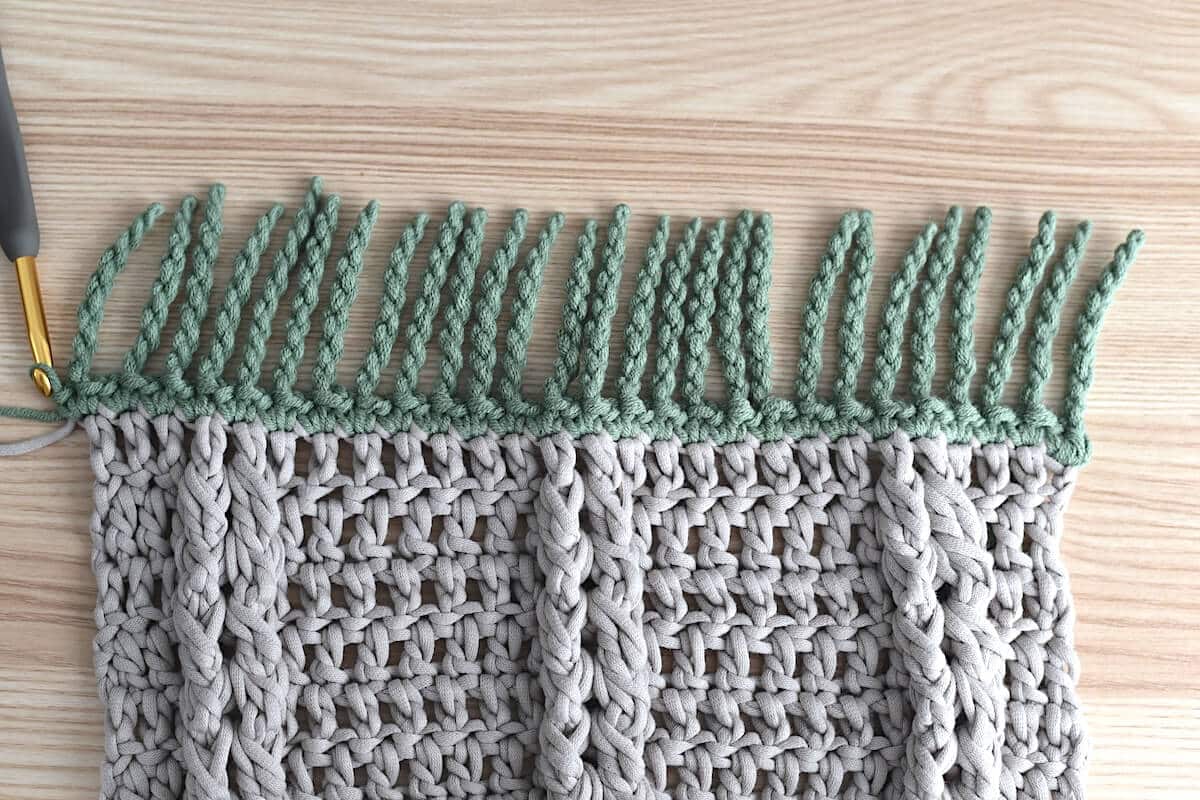 A close-up of a brown crochet swatch, showing a twisted fringe made with green yarn for crochet blankets.