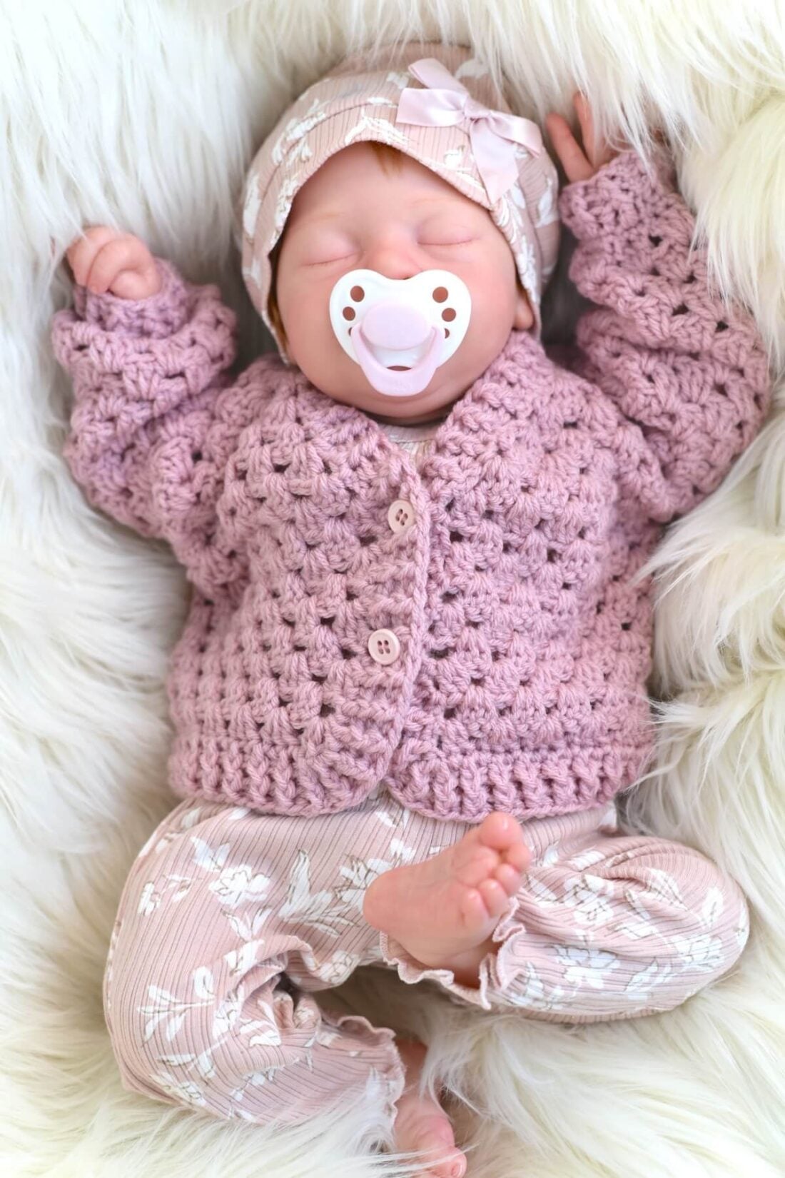 An adorable, pink crochet baby cardigan displayed on a life-size baby doll.