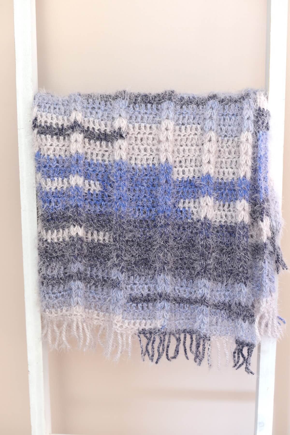 Folded crochet throw blanket with ribbing texture and fringe edges in gray, cream and blue tones.