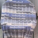 Crochet blanket draped over a chair, featuring ribbed texture and color blocks of gray, blue, and white.