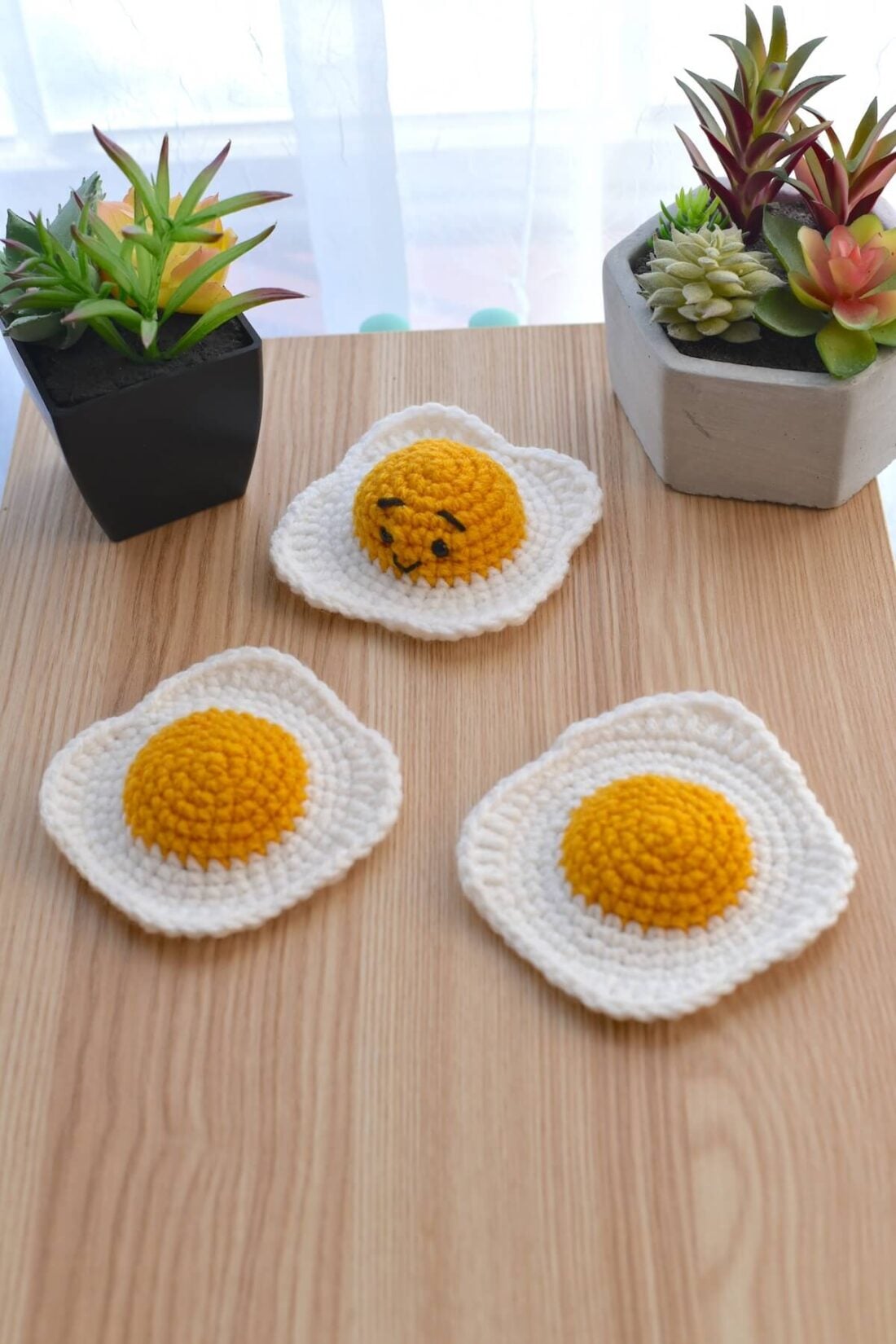 Crochet fried egg plushes created with acrylic yarn on a wooden surface and cactus plants in the background.