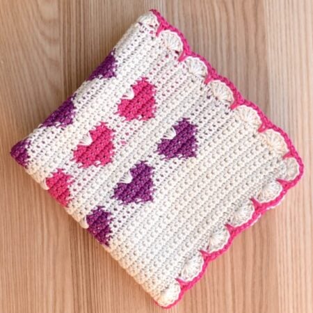 A folded Valentine’s Day square crochet dishcloth with heart colorwork design and pink edge.