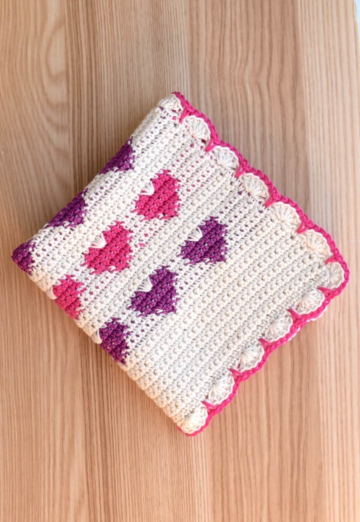 A folded Valentine’s Day square crochet dishcloth with heart colorwork design and pink edge.