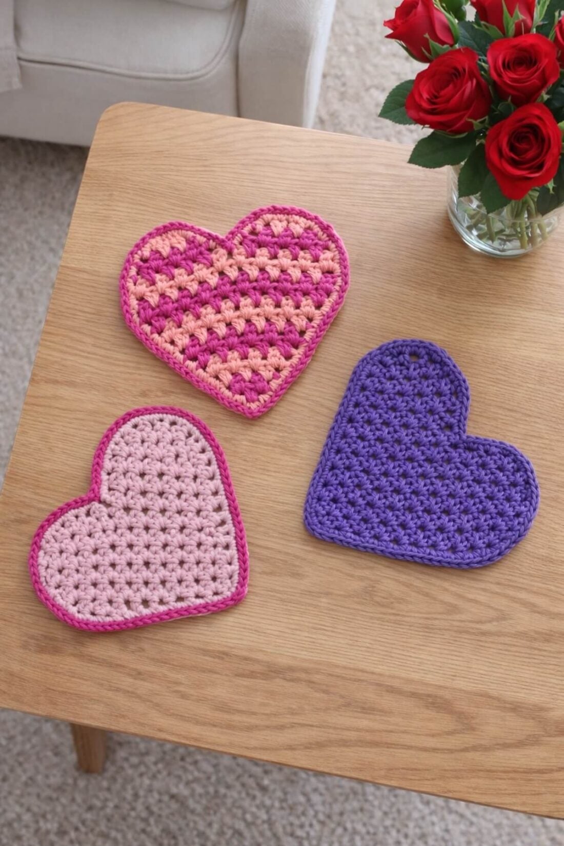 Three heart-shaped crochet beermats are displayed on a coffee table with a vase of roses in the background.
