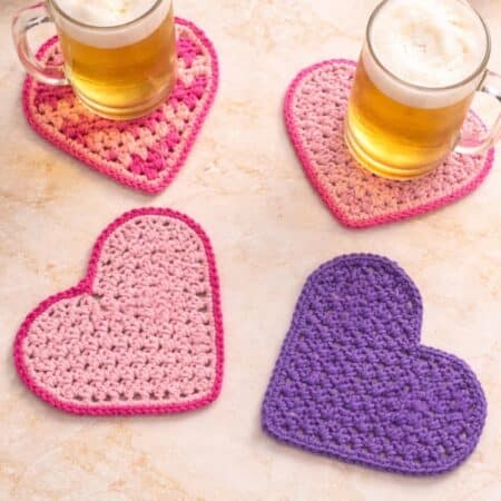 Four heart shaped crochet beermats displayed on a on a champagne marbled kitchen counter with two beer mugs on it.