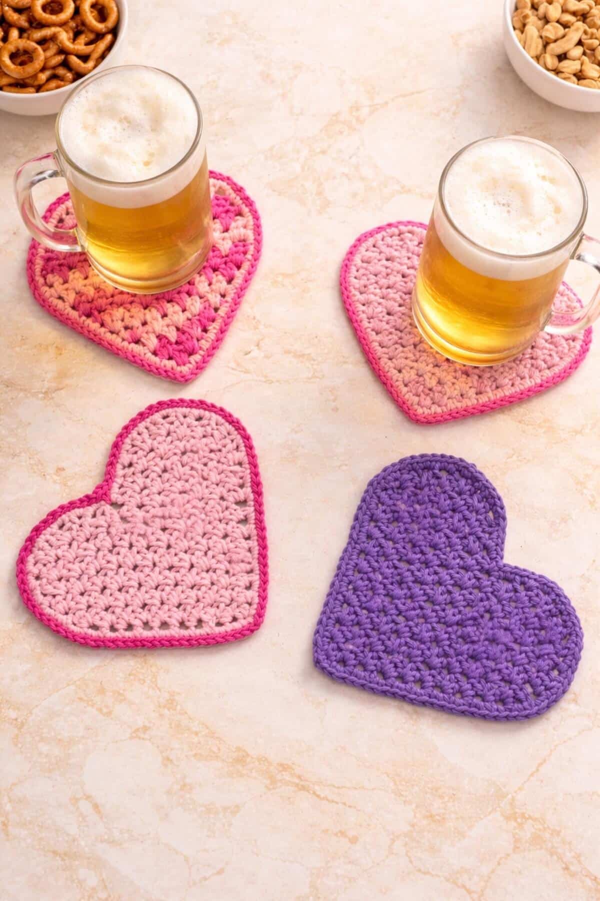 Four heart shaped crochet beermats displayed on a on a champagne marbled kitchen counter with two beer mugs on it.