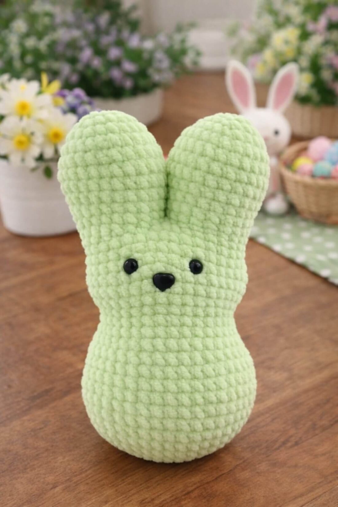 Crochet marshmallow peep bunny Amigurumi in soft green color sitting on a wooden table with Easter decorations in the background.
