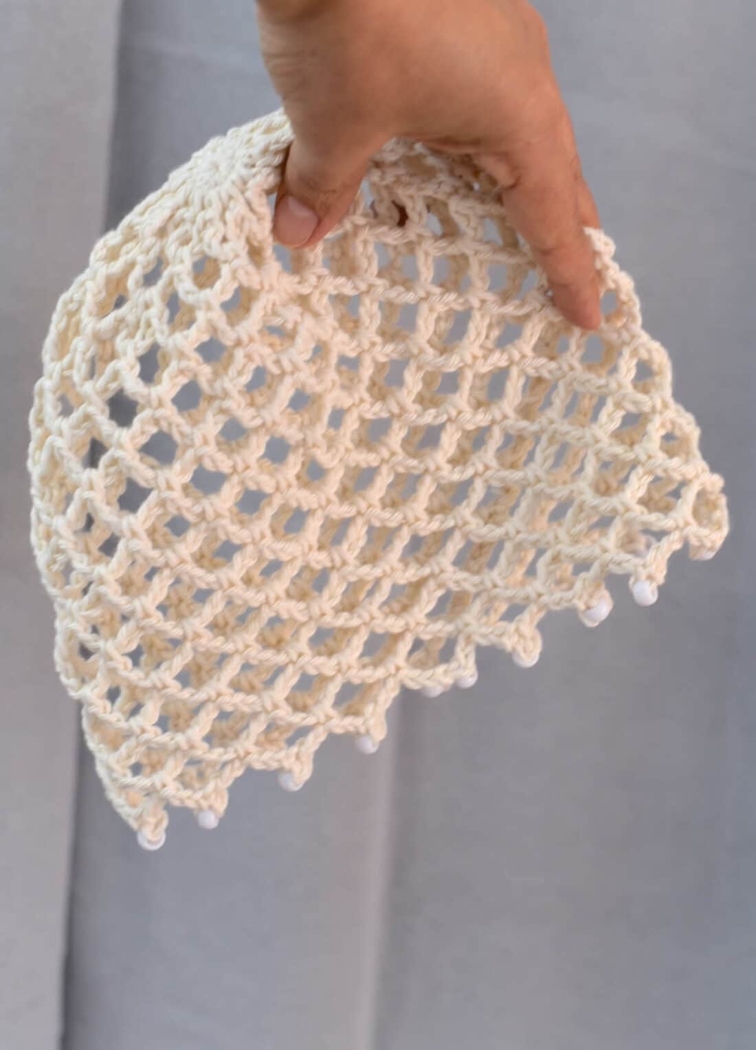 A hand holding a crochet skull cap pattern in cotton yarn with open stitch design