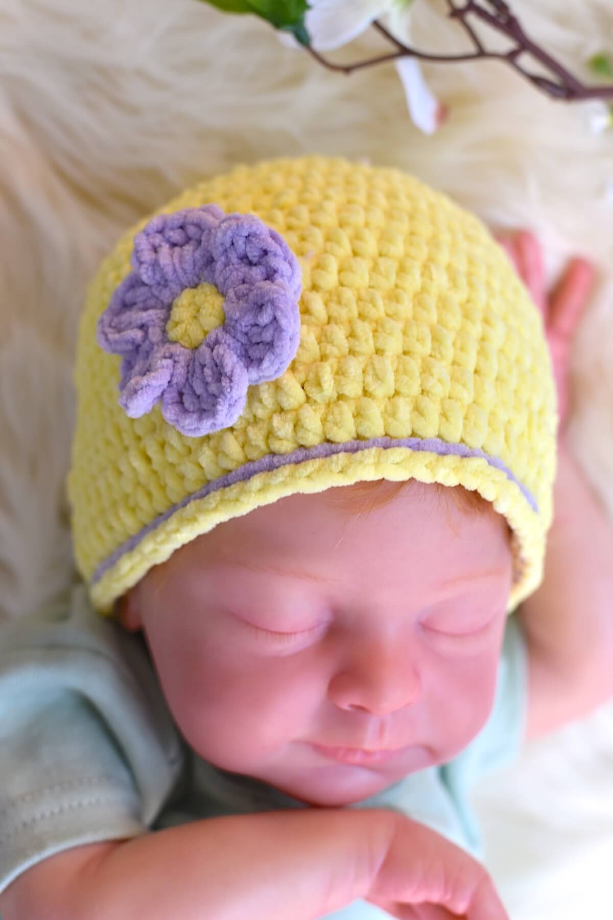 Sleeping baby modeling a handmade crochet newborn hat with decorative purple flower and purple trim.