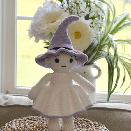 Crochet witch doll wearing a purple pointed hat and white skirt, standing on the bottom of a basket.