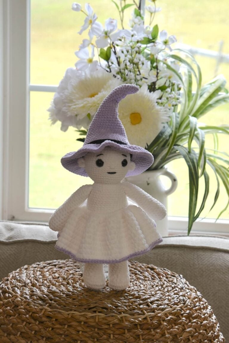 Crochet witch doll wearing a purple pointed hat and white skirt, standing on the bottom of a basket.